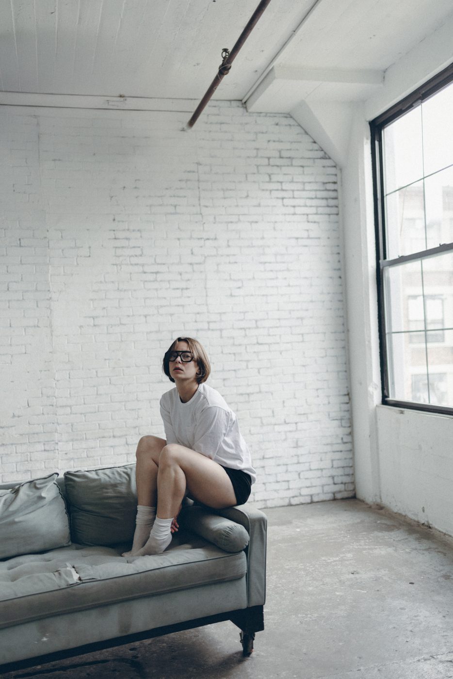 Woman in glasses and a white tee perched on a grey couch against a white brick wall