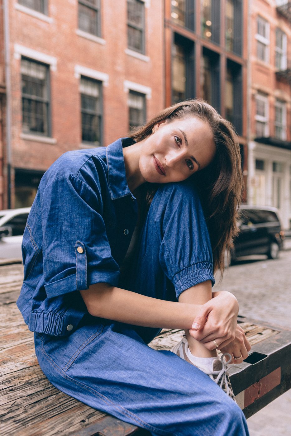 Woman in a denim set resting her head on her knee, brick townhouses behind her
