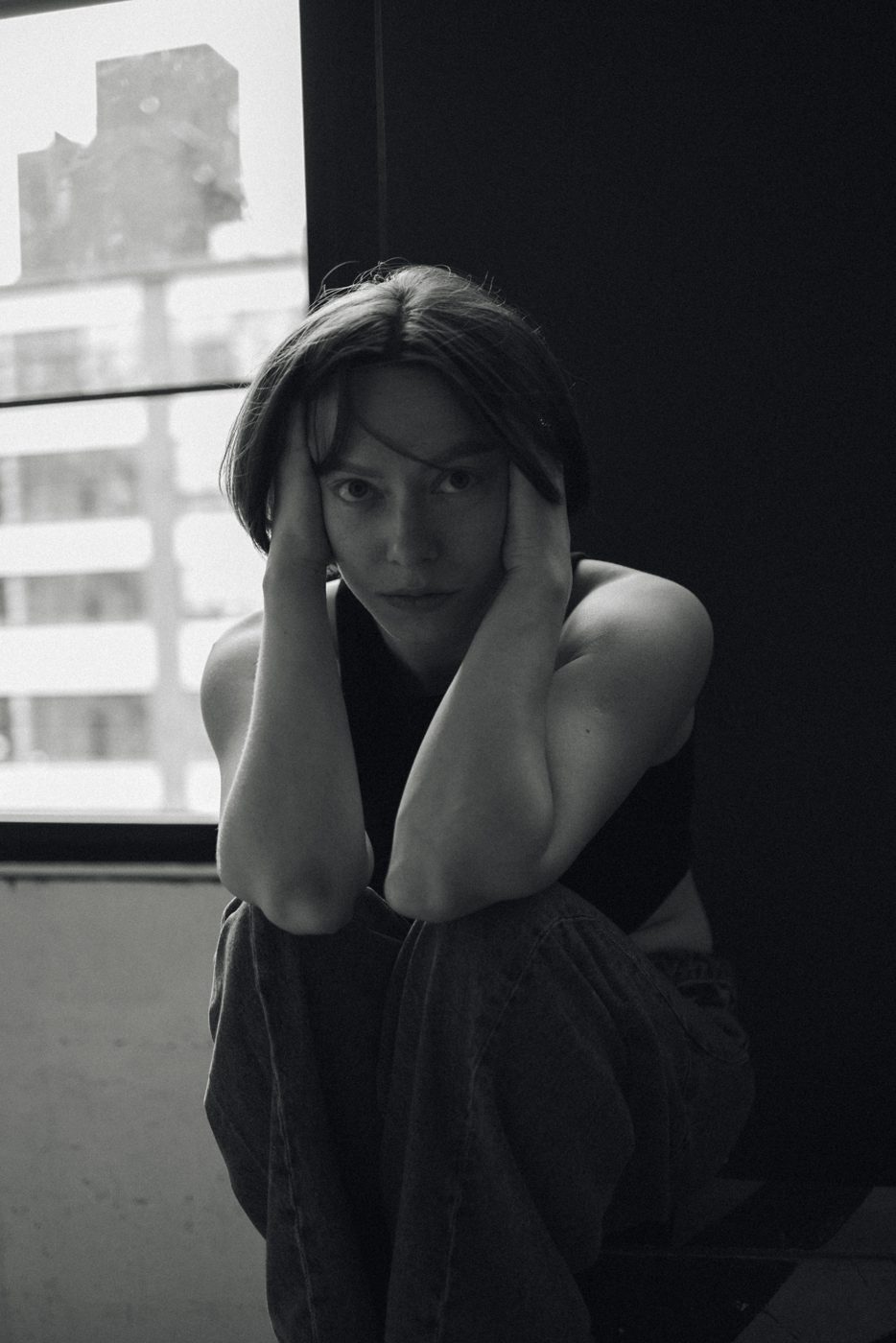 Woman crouched by a window, hands pressed to her temples, looking straight into the camera