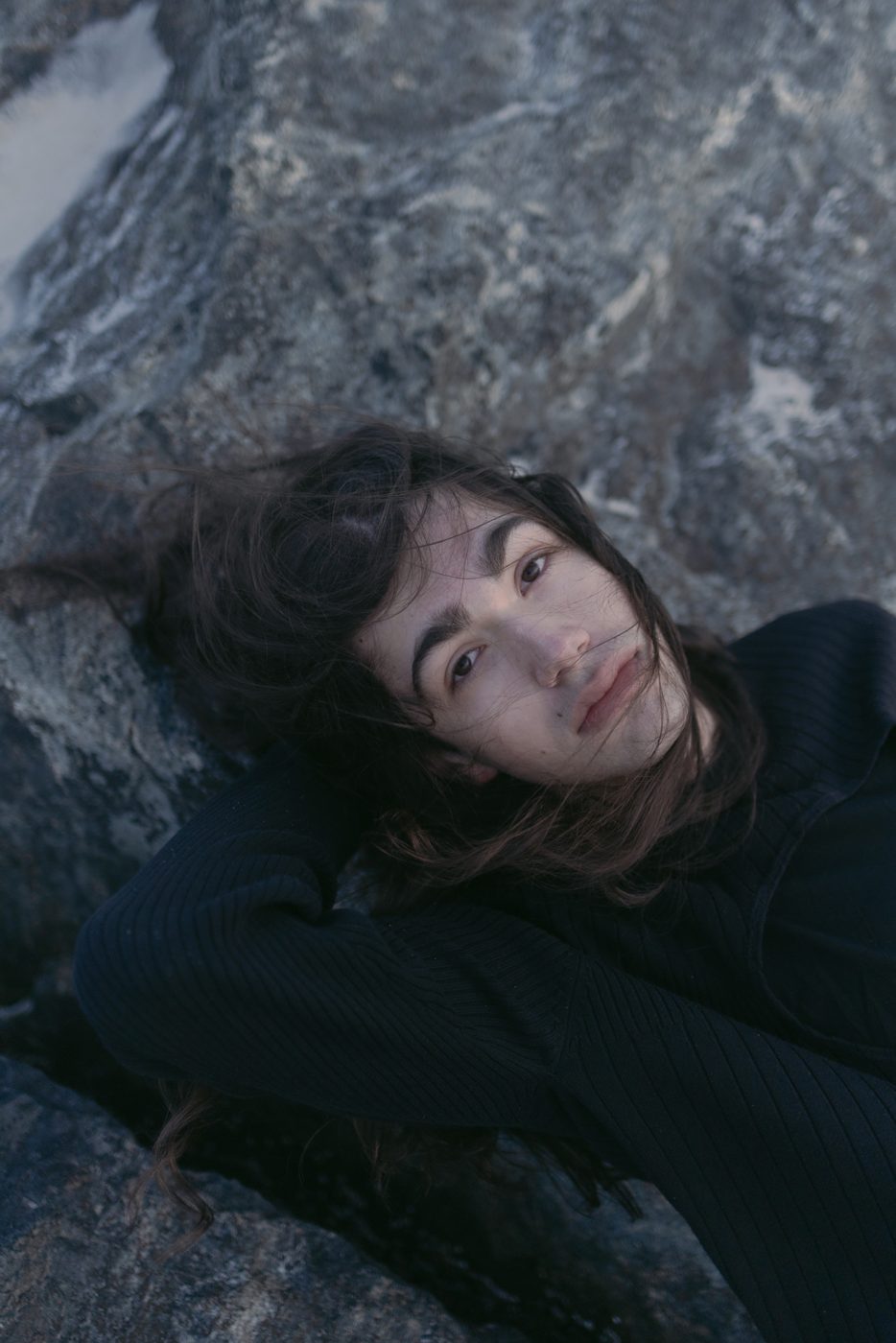 Person with long dark hair lying on grey rocks, looking up into the camera, in muted colour